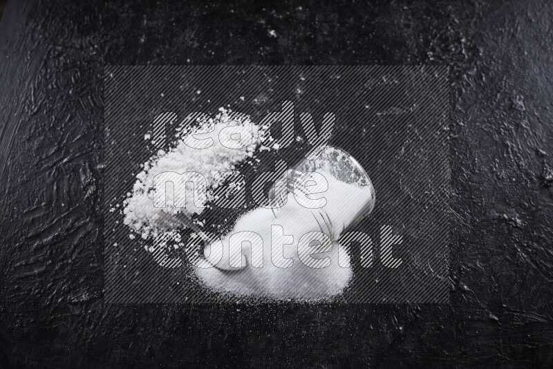 A glass jar full of table salt with some sea salt crystals beside it on a black background