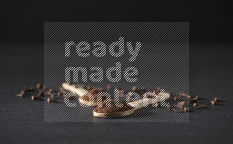 ملعقتان خشبيتان ممتلئتان ببودرة القرنفل مع حبوب القرنفل المتناثرة على خلفية سوداء