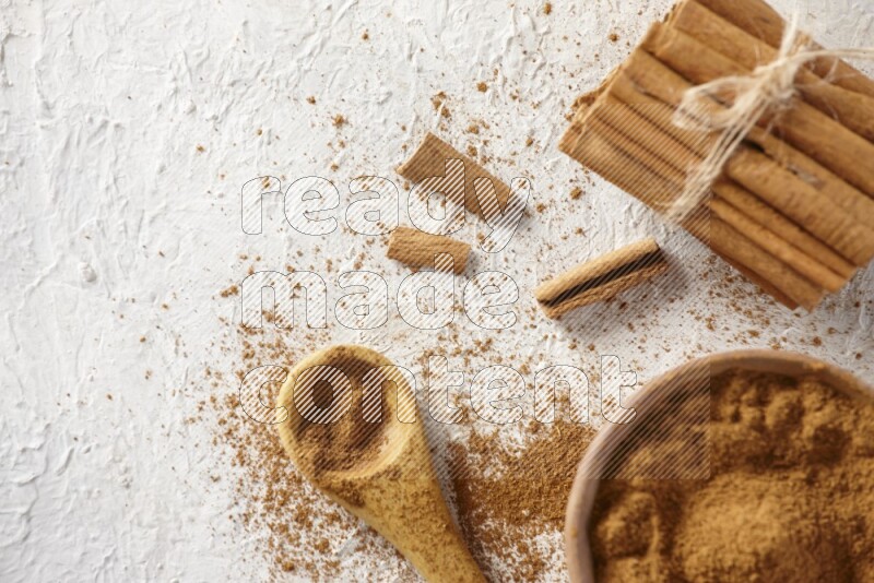 Cinnamon sticks stacked and bounded beside a wooden bowl full of cinnamon powder and a wooden spoon full of powder on white background