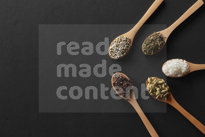 wooden spoons filled with white peppers, cloves, cardamom, salt, black peppers and basil on black flooring