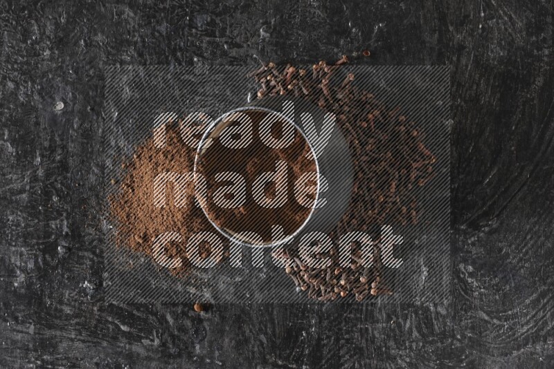A black pottery bowl full of cloves powder spilled out of it with whole grains on a textured black flooring
