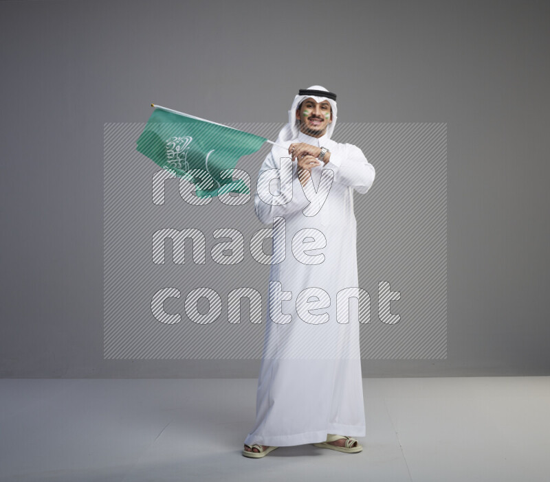 A Saudi man standing wearing thob and white shomag with face painting raising big Saudi flag on gray background