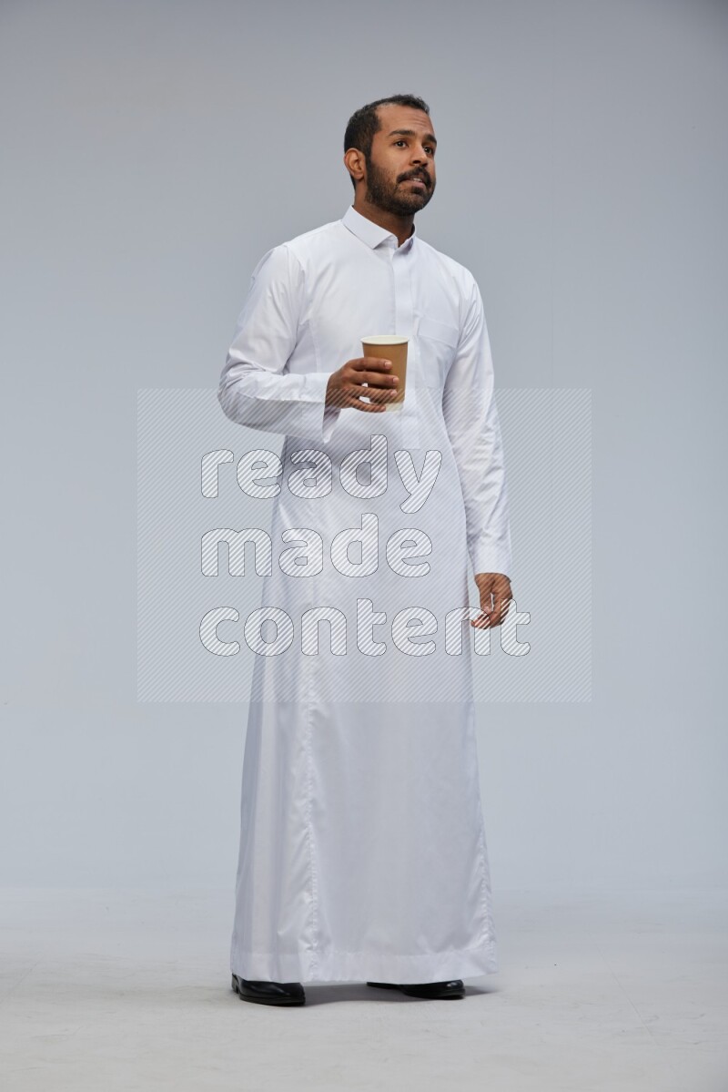 Saudi man wearing thob standing holding paper cup on gray background