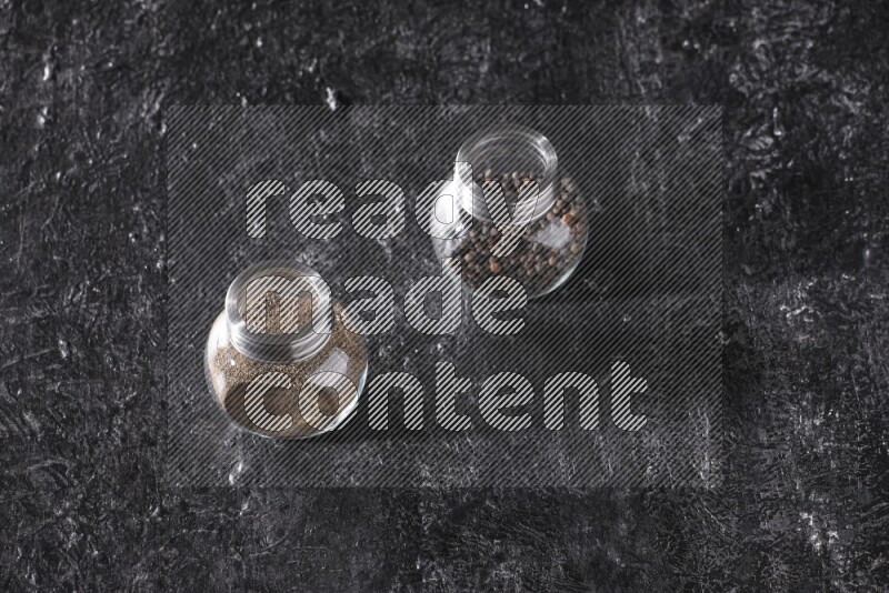 2 glass spice jars full of black pepper powder and black pepper beads on textured black flooring