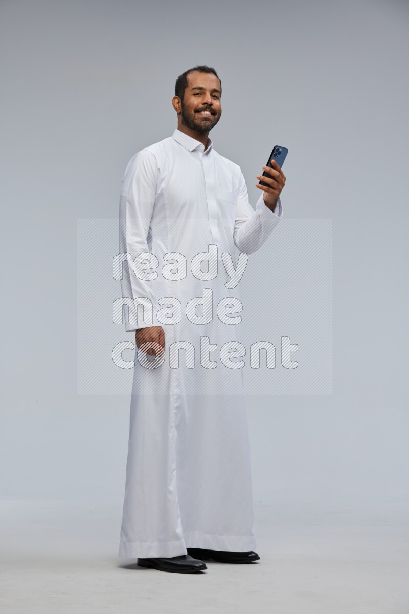 Saudi man wearing Thob standing texting on phone on Gray background