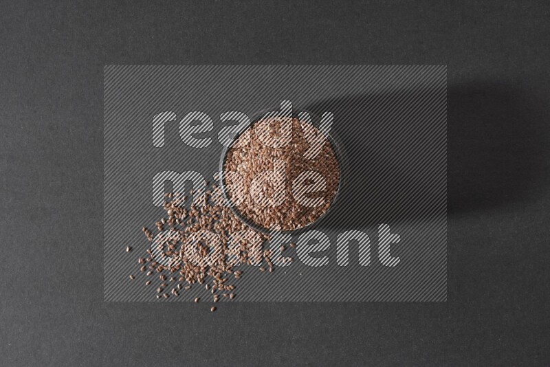 A black pottery bowl full of flaxseeds surrounded by the seeds on a black flooring
