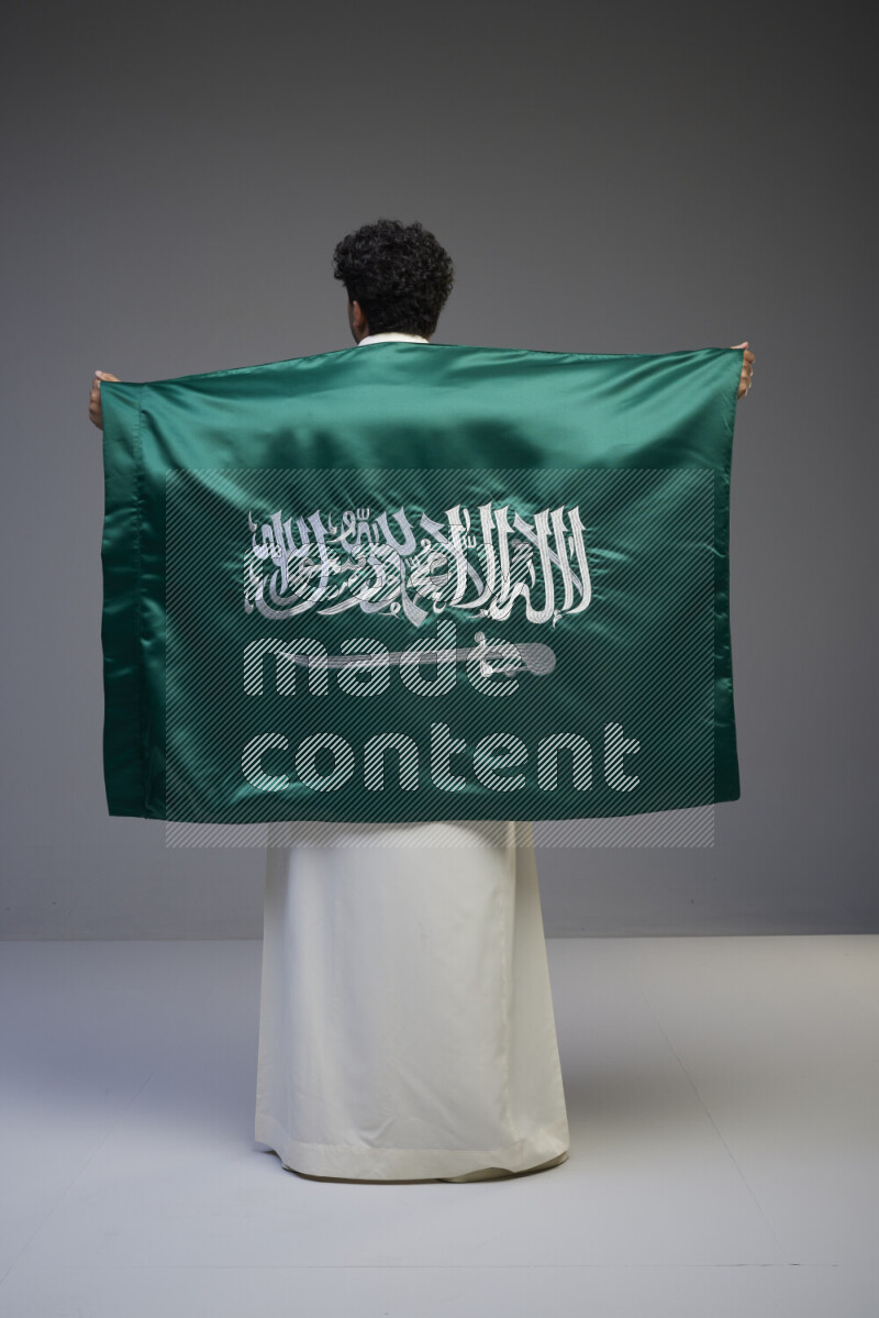 A Saudi man standing wearing thob holding big Saudi flag on gray background