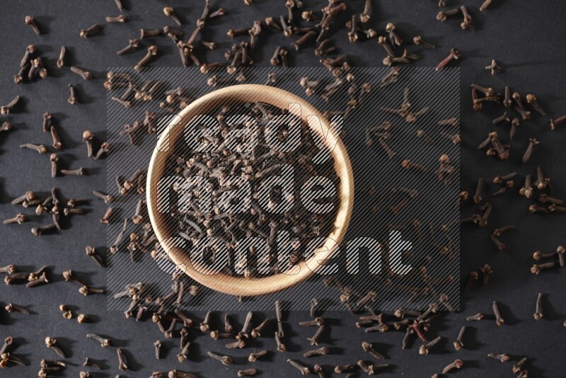 A wooden bowl full of cloves with spreaded whole cloves on a black flooring