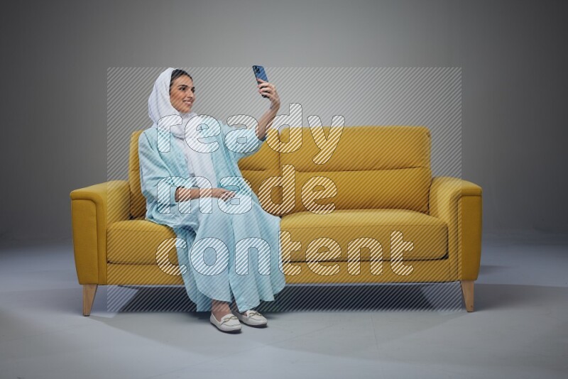 A Saudi woman wearing a light blue Abaya and white head scarf sitting on a yellow sofa and taking selfies with her phone eye level on a grey background