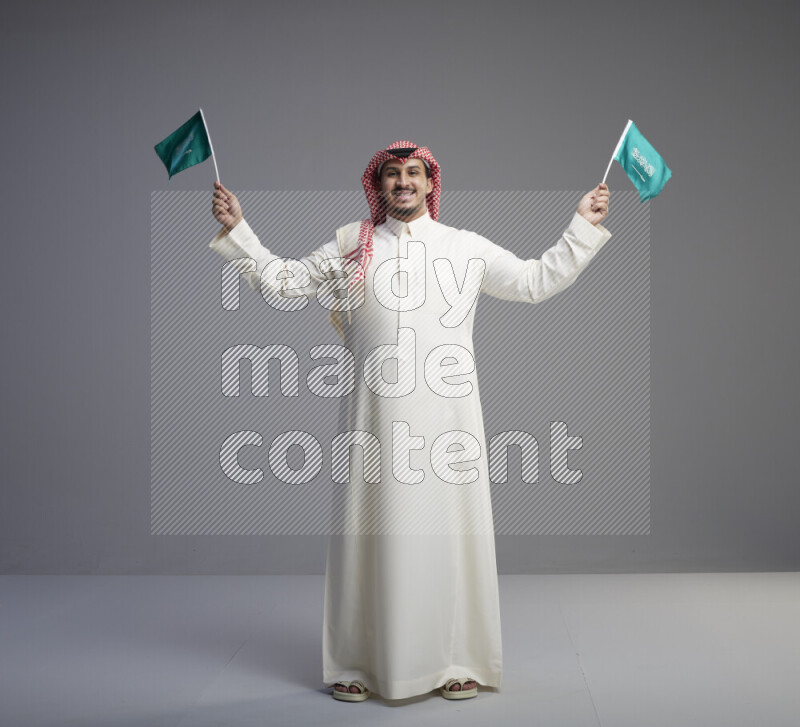A Saudi man standing wearing thob and red shomag raising small Saudi flag on gray background