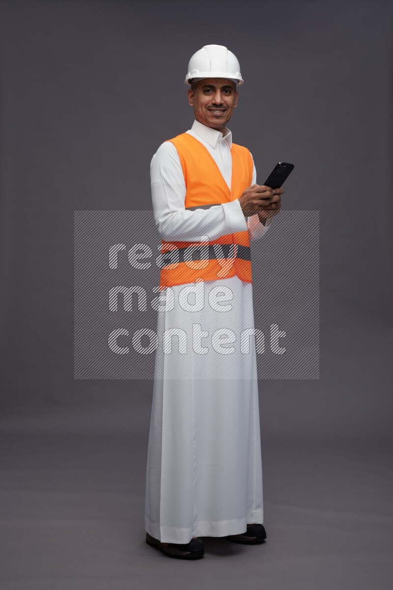 Saudi man wearing thob with engineer vest standing texting on phone on gray background