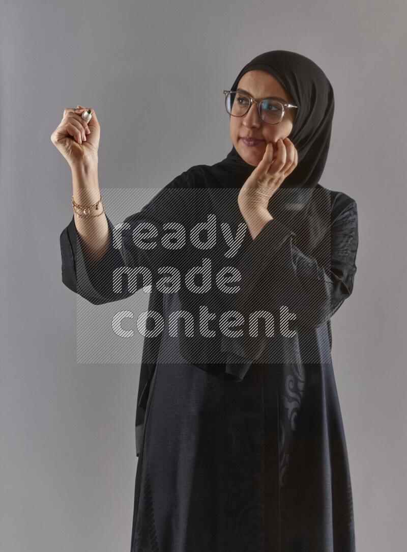 Woman in black abaya, hijab and glasses holding a marker pen to write on a transparent board with different reactions and expressions on grey background