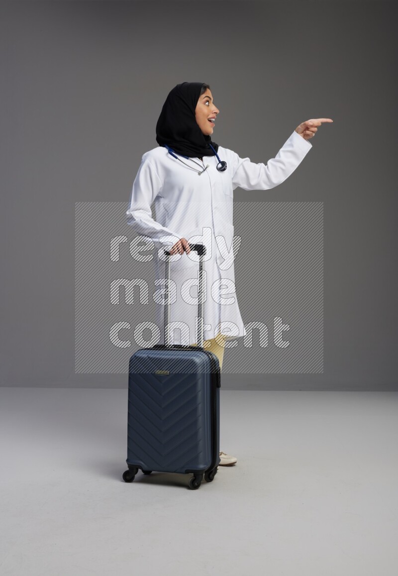 Saudi woman wearing lab coat with stethoscope standing holding Travel bag on Gray background
