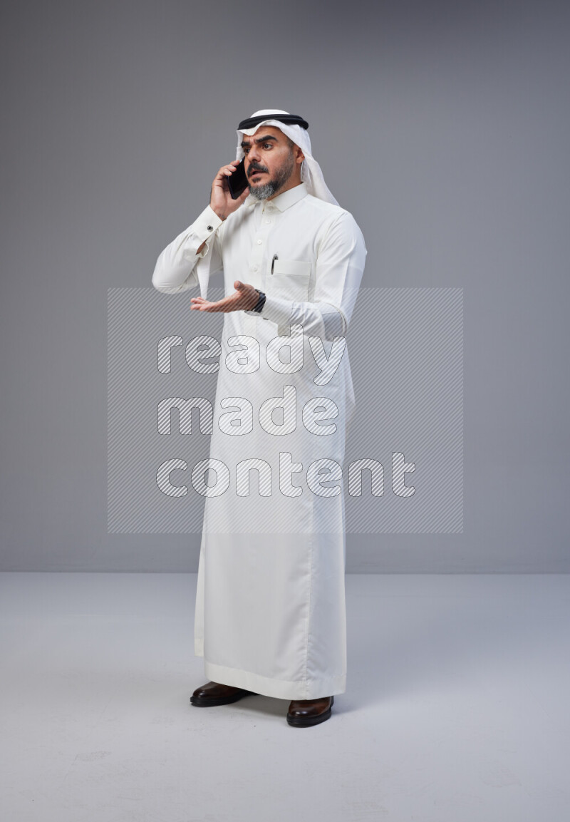 Saudi man Wearing Thob and white Shomag standing talking on phone on Gray background