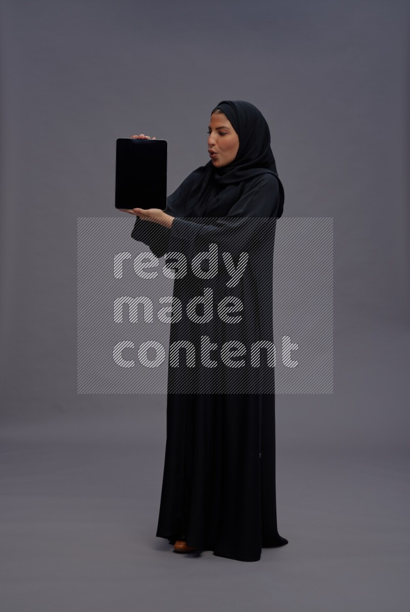 Saudi woman wearing Abaya standing showing tablet to camera on gray background