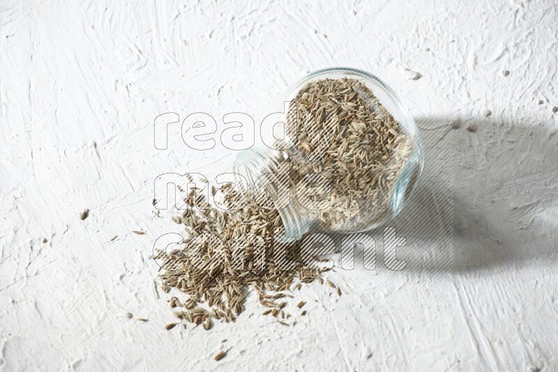 A flipped glass spice jar full of cumin seeds and the seeds spilled out on textured white flooring