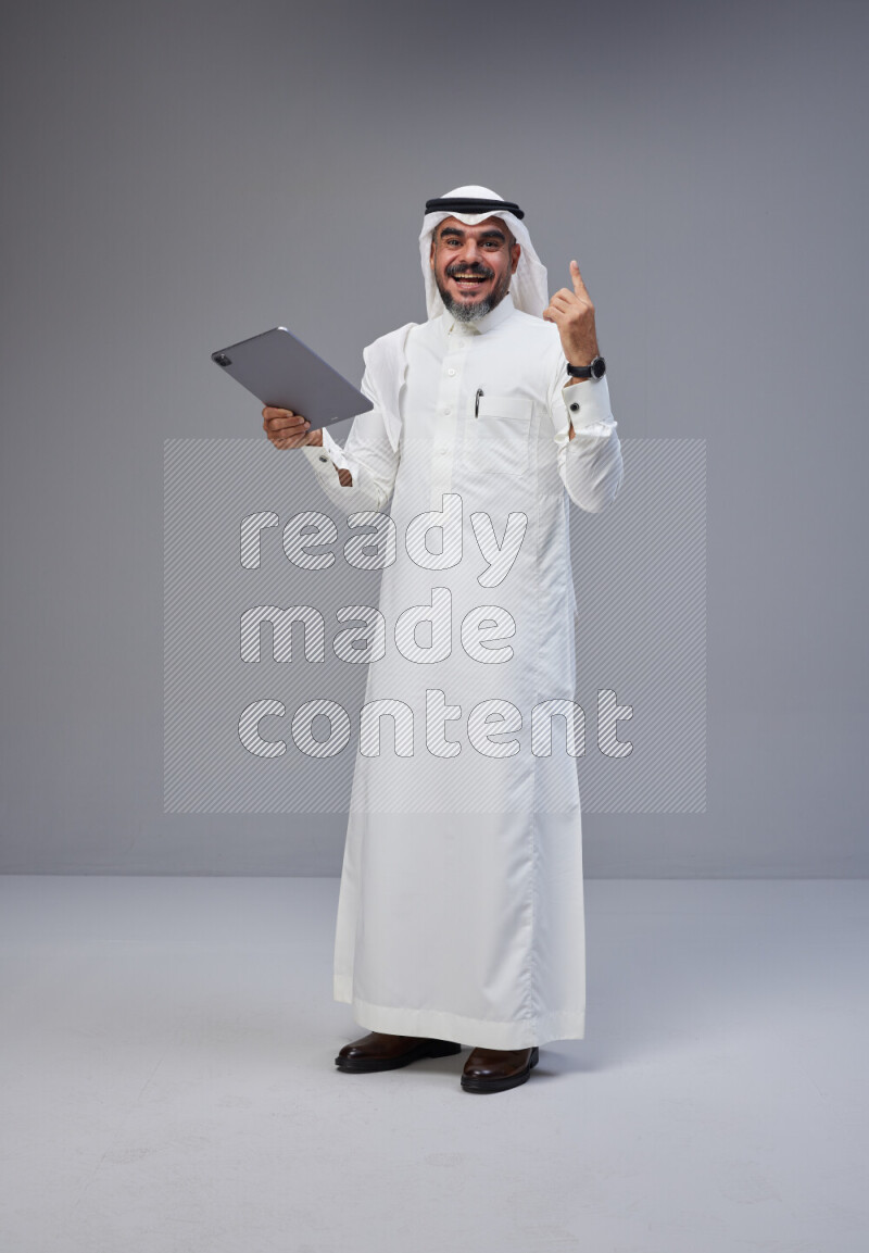 Saudi man Wearing Thob and white Shomag standing working on tablet on Gray background