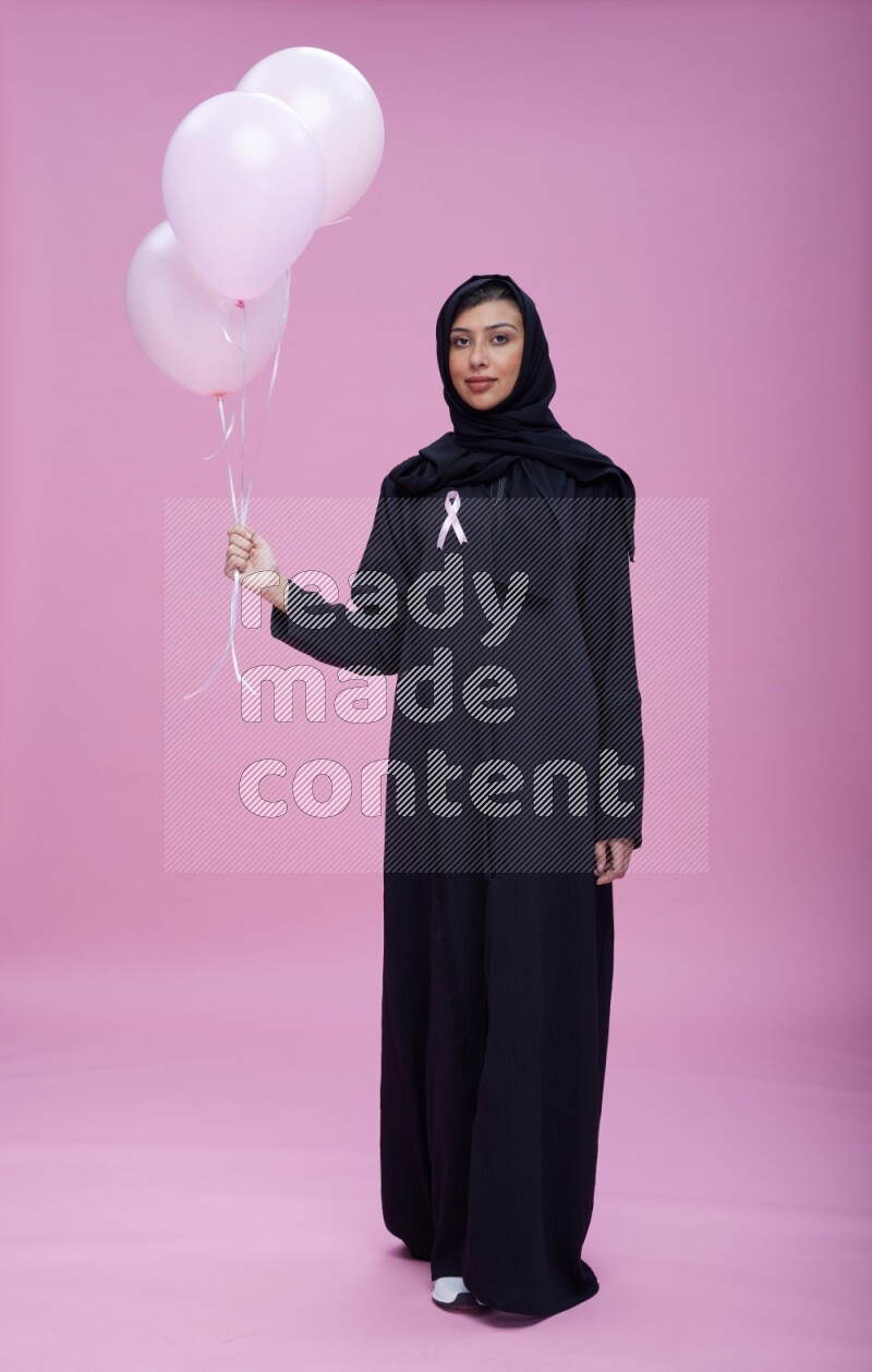 Saudi woman wearing pink ribbon on Abaya standing holding balloons on plain pink background