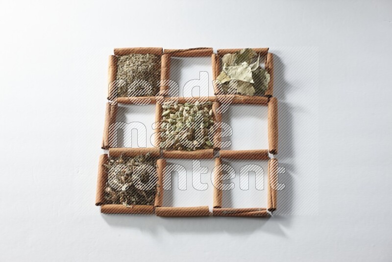 9 squares of cinnamon sticks full of cardamom in the middle surrounded by nutmeg, cinnamon, bay laurel leaves, cloves, cumin, dried ginger, dried basil and star anise on white flooring