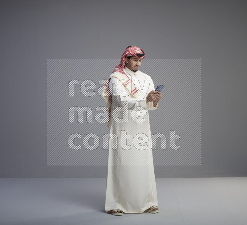 A  Saudi man standing wearing thob and red shomag texting on phone on gray background