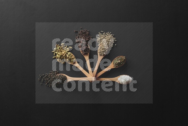 الفلفل الأبيض والقرنفل والهيل والملح والفلفل الأسود وريحان مجفف في معالق خشبية علي خلفية سوداء