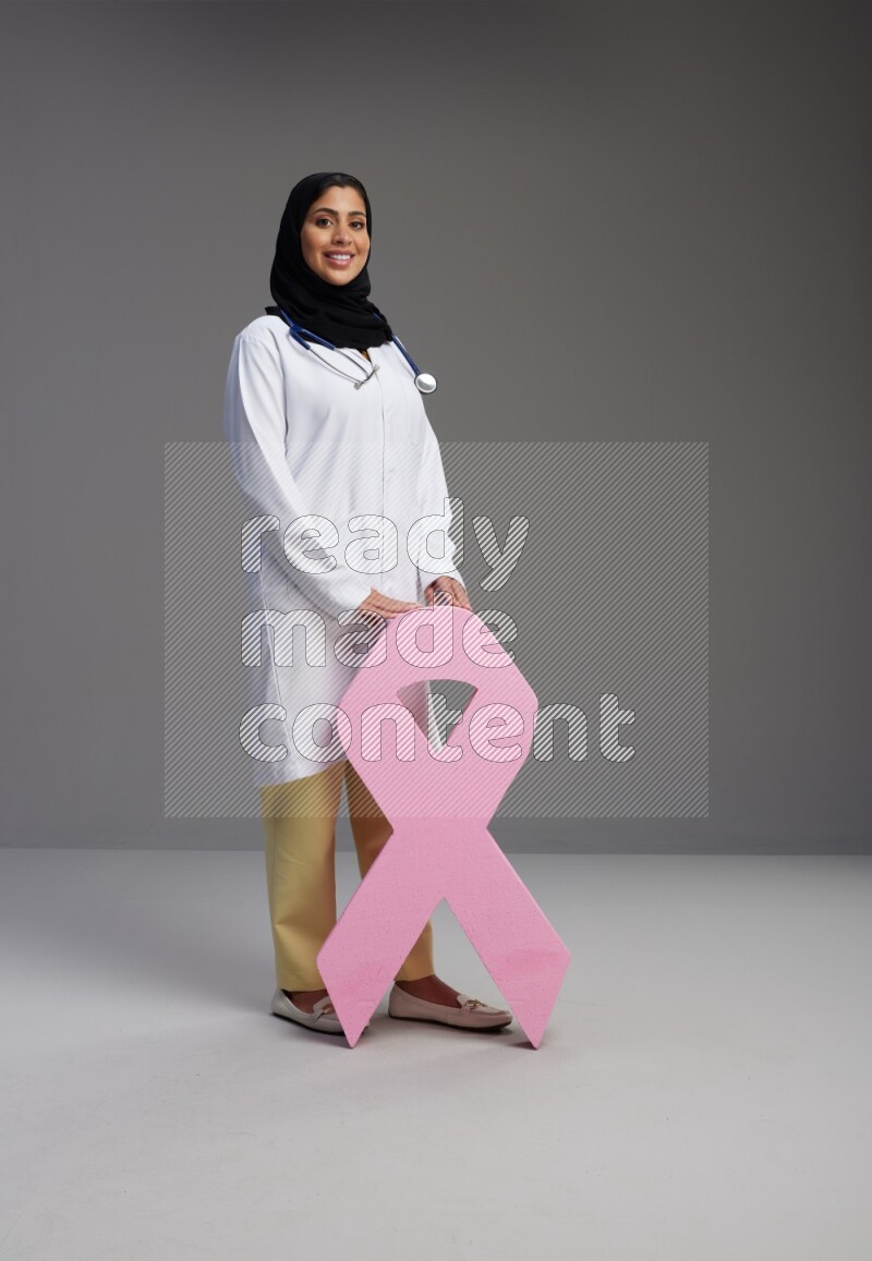 Saudi woman wearing lab coat with stethoscope standing holding awareness ribbon symbol standing on Gray background