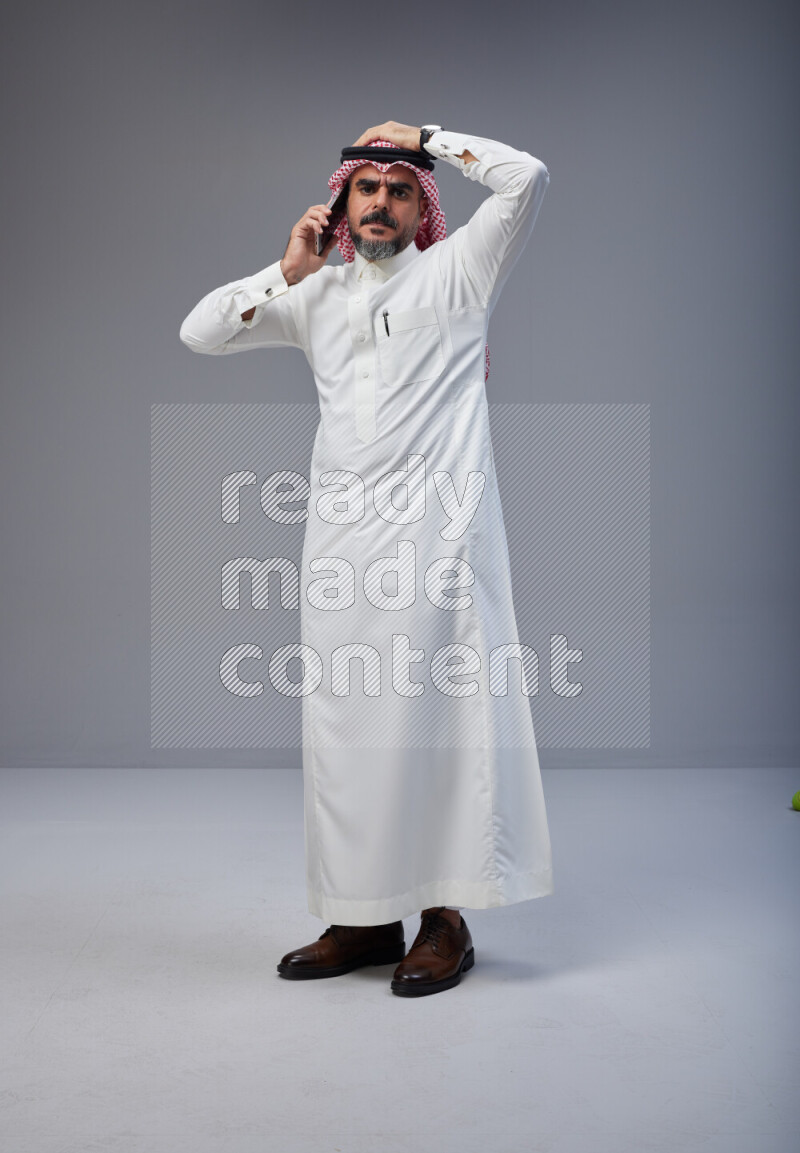 Saudi man Wearing Thob and red Shomag standing talking on phone on Gray background