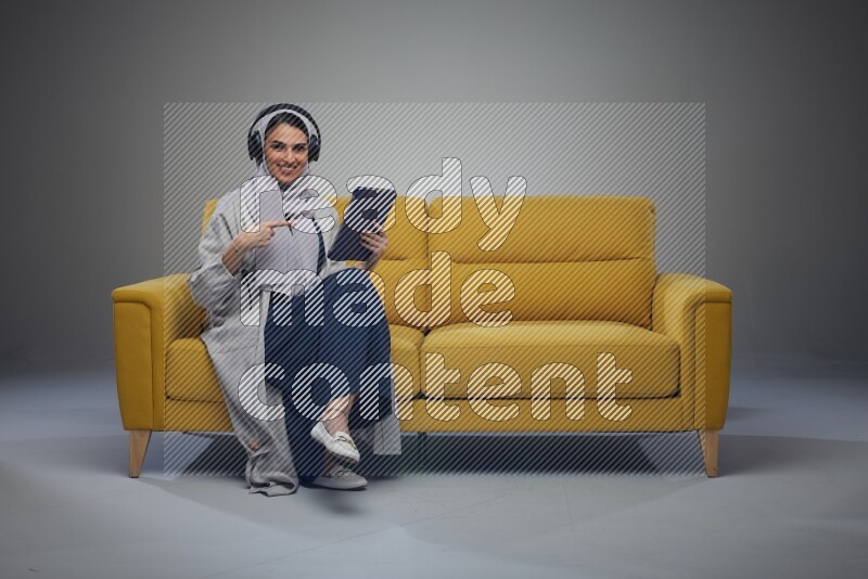 A Saudi woman wearing a light gray Abaya and head scarf sitting on a yellow sofa and using her phone while wearing headphones eye level on a grey background