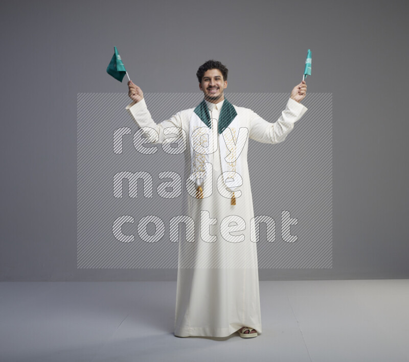 A Saudi man standing wearing thob and saudi flag scarf and holding small saudi flag on gray background