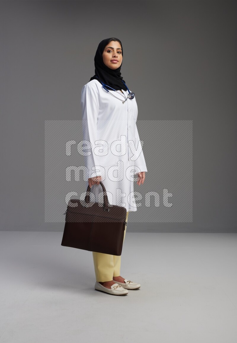 Saudi woman wearing lab coat with stethoscope standing holding bag on Gray background