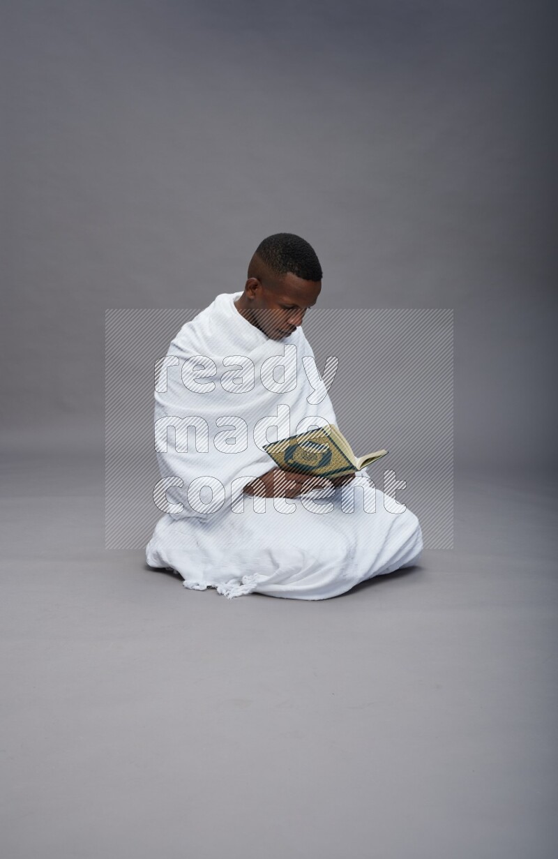 A man wearing Ehram sitting on floor reading quran on gray background