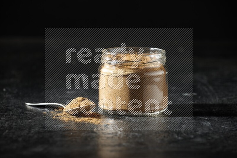 A glass jar and a metal spoon full of allspice powder on a textured black flooring