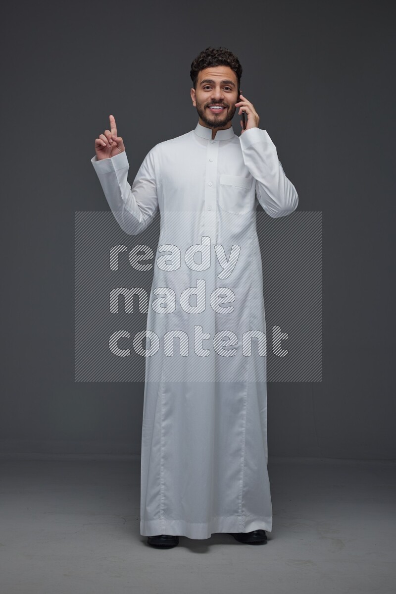 A Saudi man wearing Thobe and talking in the phone while standing and making different hand gestures eye level on a gray background