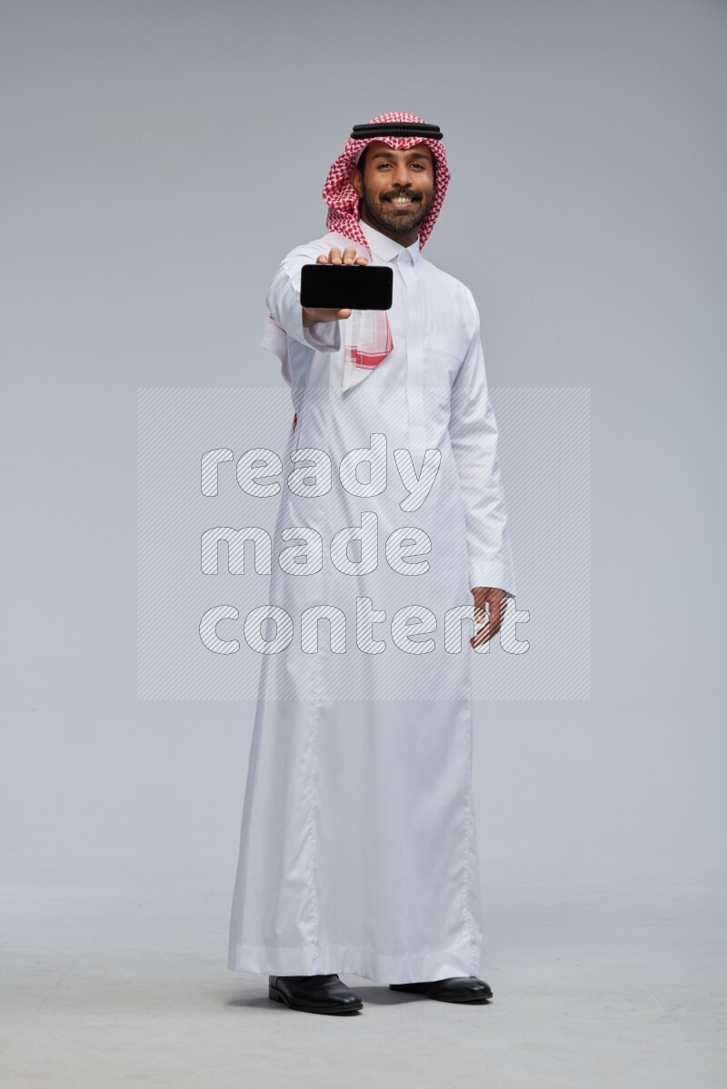 Saudi man Wearing Thob and shomag standing showing phone to camera on Gray background