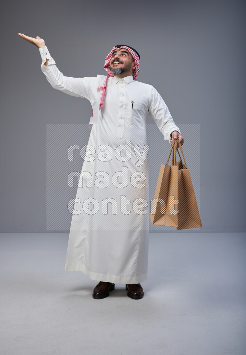 Saudi man Wearing Thob and red Shomag standing holding shopping bag on Gray background