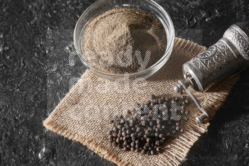 A glass bowl full of black pepper powder and black pepper beads on burlap fabric with a turkish metal pepper grinder on textured black flooring
