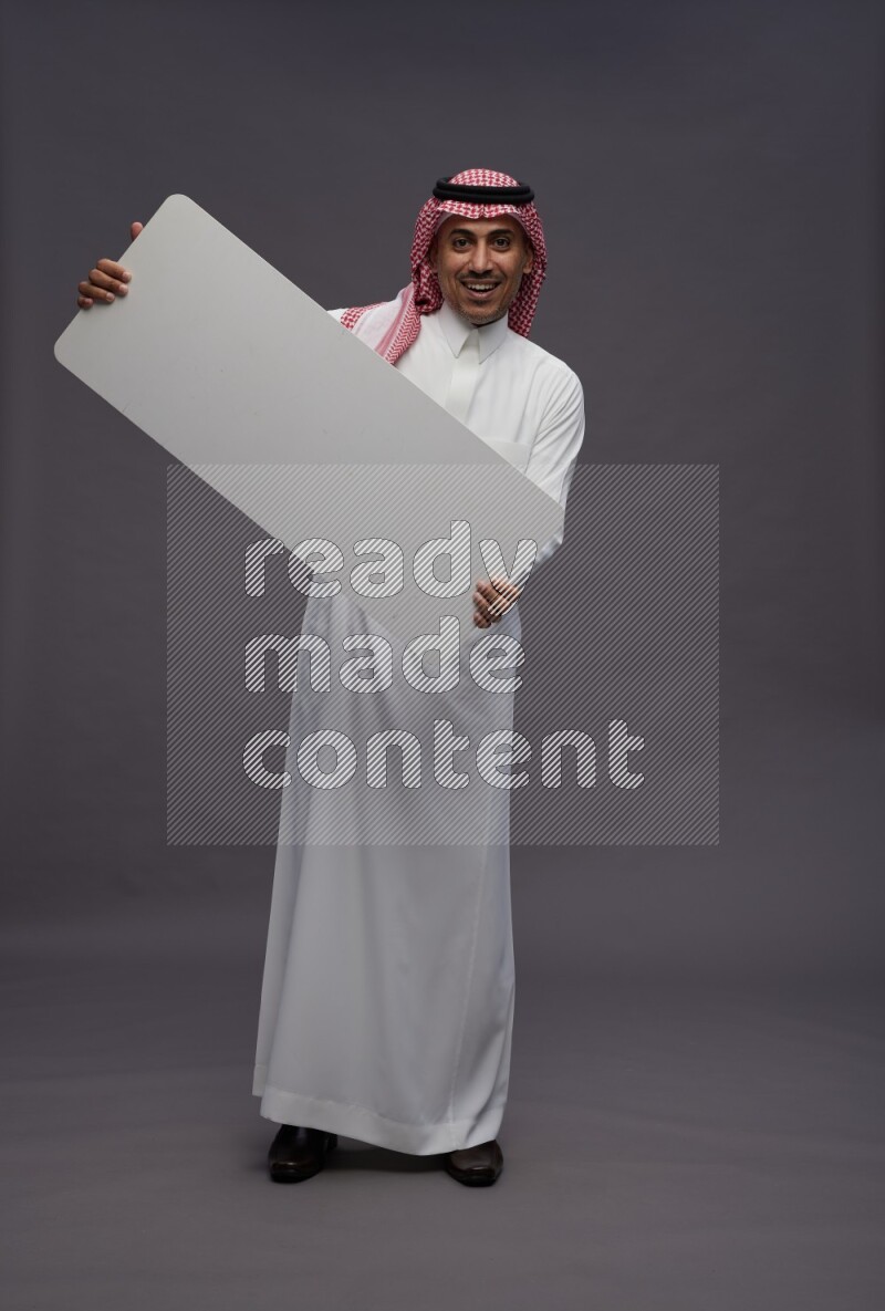 Saudi man wearing thob and shomag standing holding board on gray background