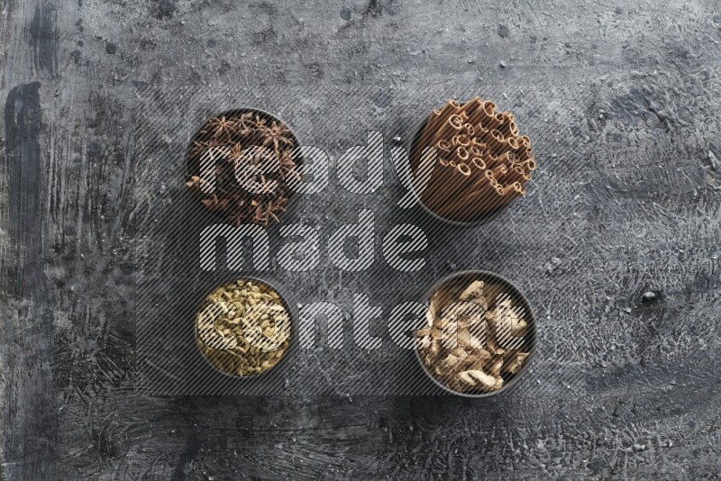 Ginger, Cardamom, Star anise and cinnamon sticks in 4 bowls on a textured black background
