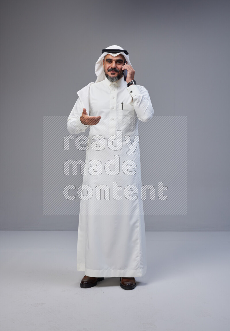 Saudi man Wearing Thob and white Shomag standing talking on phone on Gray background
