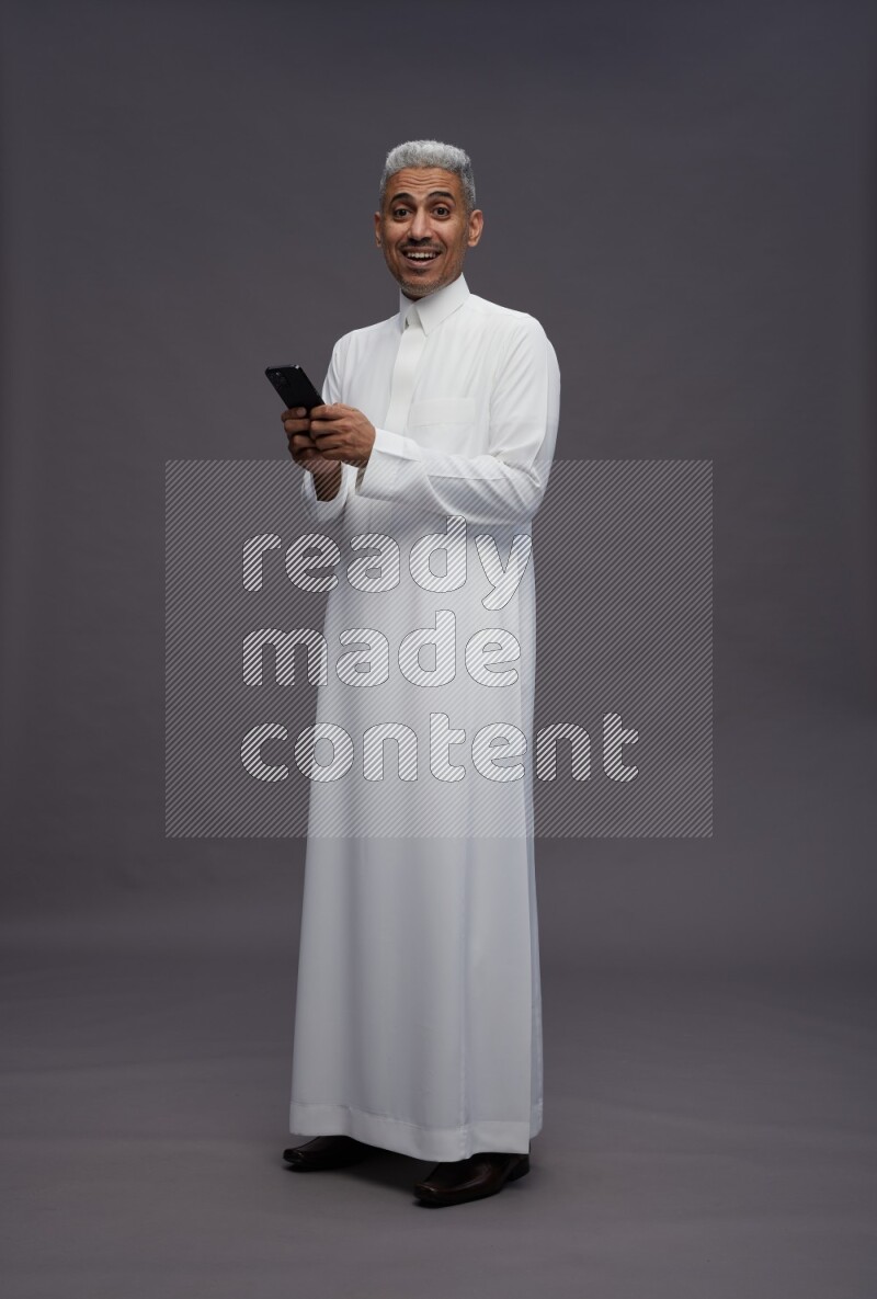 Saudi man wearing thob standing texting on phone on gray background
