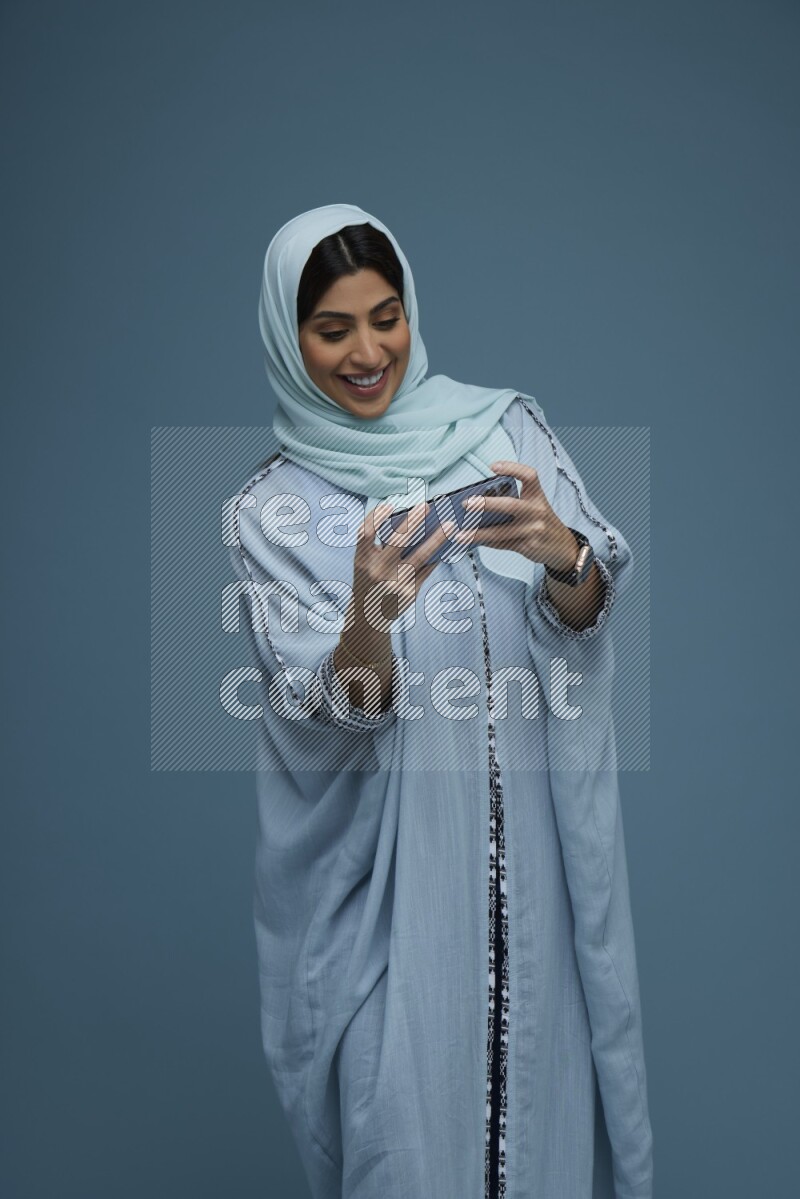 A Saudi woman Playing a Game on her phone on a blue background wearing a blue Abaya with hijab
