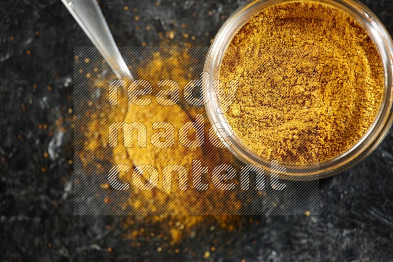 A glass jar and a metal spoon full of turmeric powder on a textured black flooring