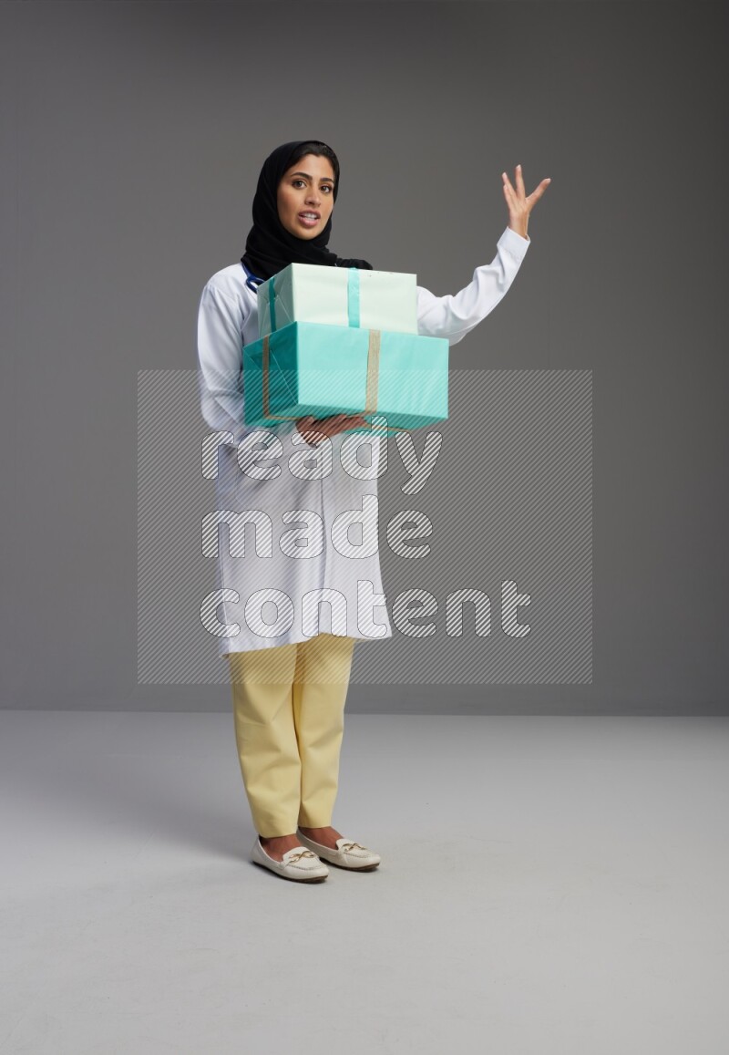Saudi woman wearing lab coat with stethoscope standing holding gift box on Gray background