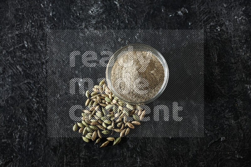 A glass bowl full of cardamom powder and cardamom seeds beside it on textured black flooring