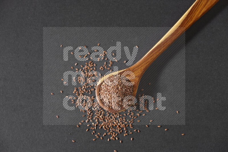 A wooden ladle full of flaxseeds and seeds spread beside it on a black flooring