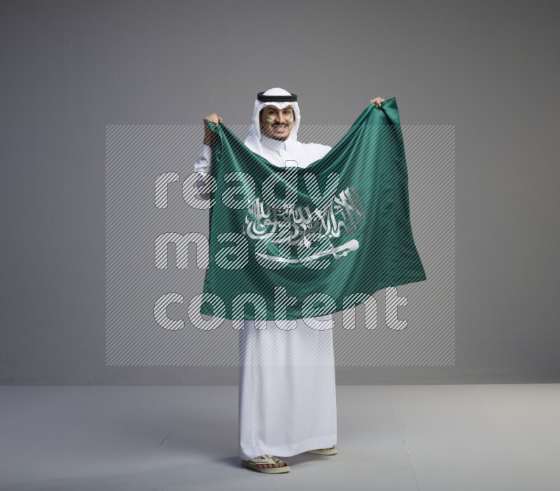 A Saudi man standing wearing thob and white shomag with face painting holding big Saudi flag on gray background