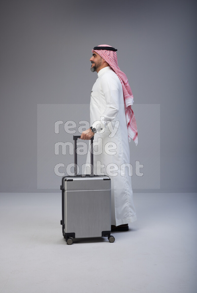 Saudi man wearing Thob and red Shomag standing holding Travel bag on Gray background