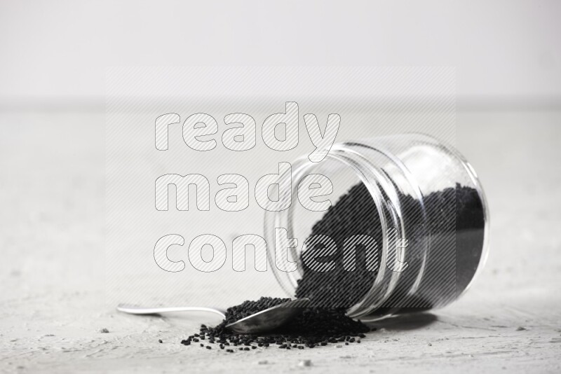 A glass jar and a metal spoon full of black seeds and the jar flipped and seeds spread on a textured white flooring