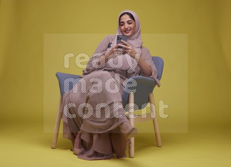 A Female Texting Sitting  on a Yellow Background wearing Brown Abaya with Hijab