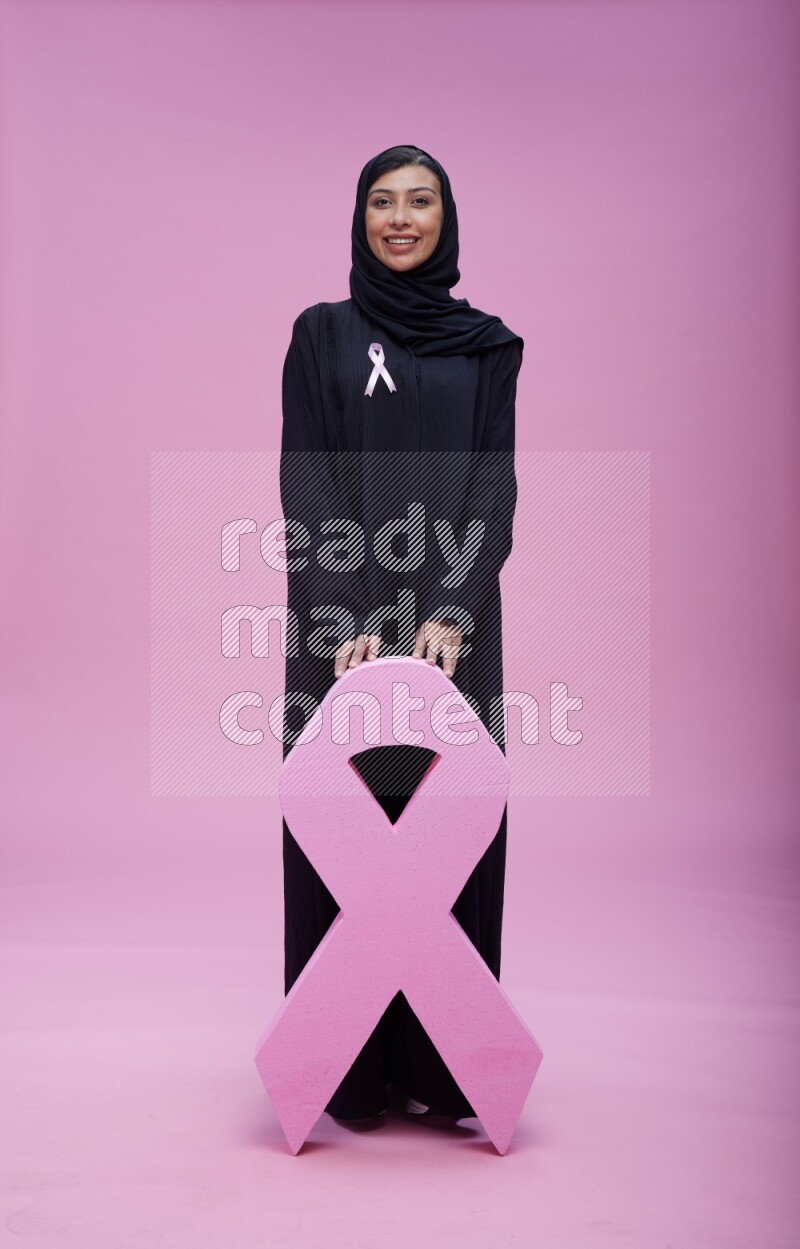 Saudi woman wearing pink ribbon on abaya standing holding awareness ribbon on plain pink background
